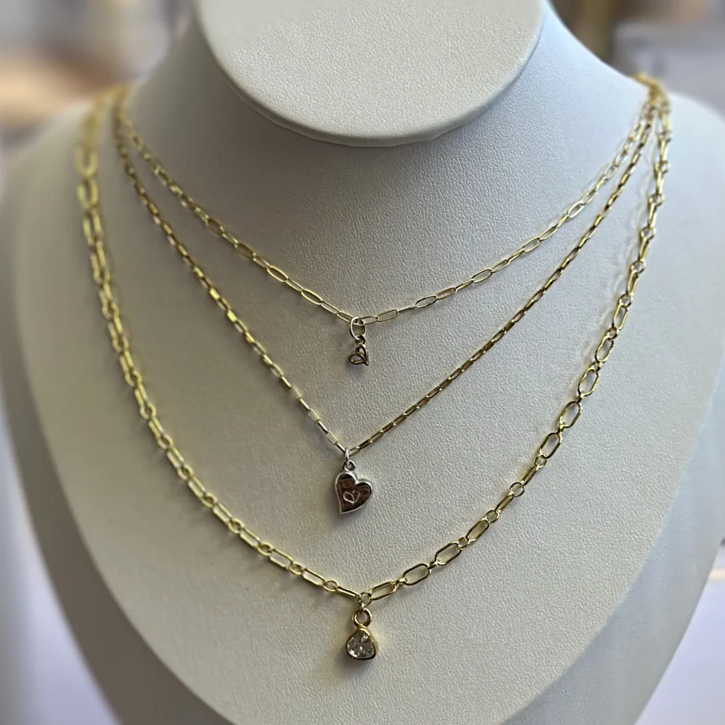 Three necklaces, gold strength chain with gold 4mm authentic pendant, gold strength chain with silver authentic heart pendant, gold courage chain with gold polki diamond on a tan jewelry bust.